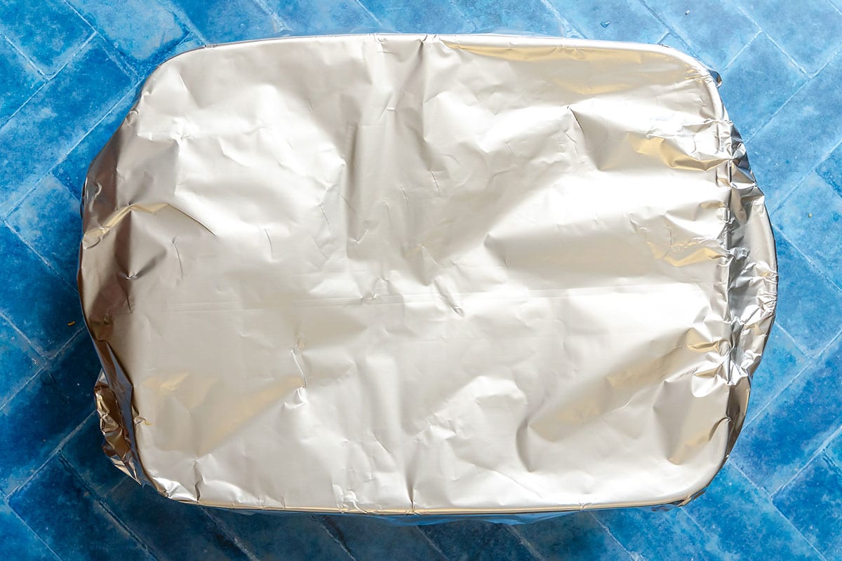 A rectangular baking dish covered with aluminum foil sits on a blue tiled surface.