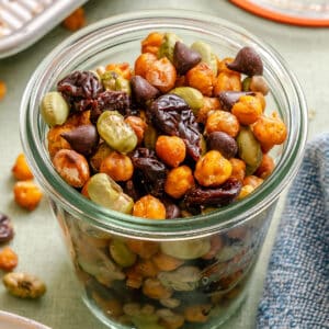 A glass jar filled with a snack mix of roasted chickpeas, fava beans, dried cherries, and chocolate chips sits on a table.