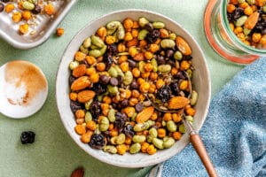 A bowl filled with roasted chickpeas, edamame, almonds, and dried cherries sits on a green tablecloth with a spoon and a blue towel nearby.
