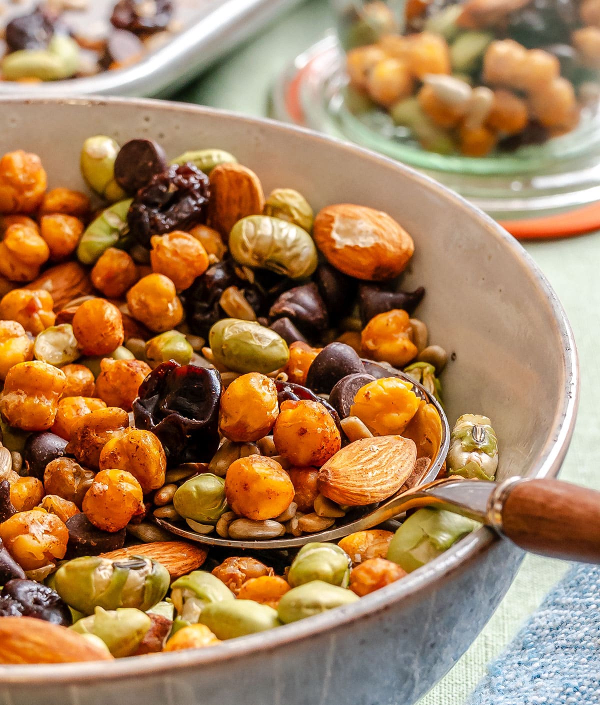 A bowl of mixed roasted chickpeas, almonds, fava beans, chocolate chips, dried berries, and seeds with a spoon inside.