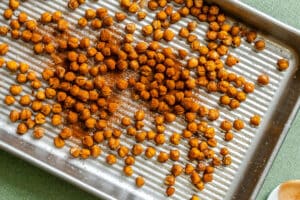 Roasted chickpeas, seasoned with spices, are spread on a ridged metal baking tray placed on a green surface.