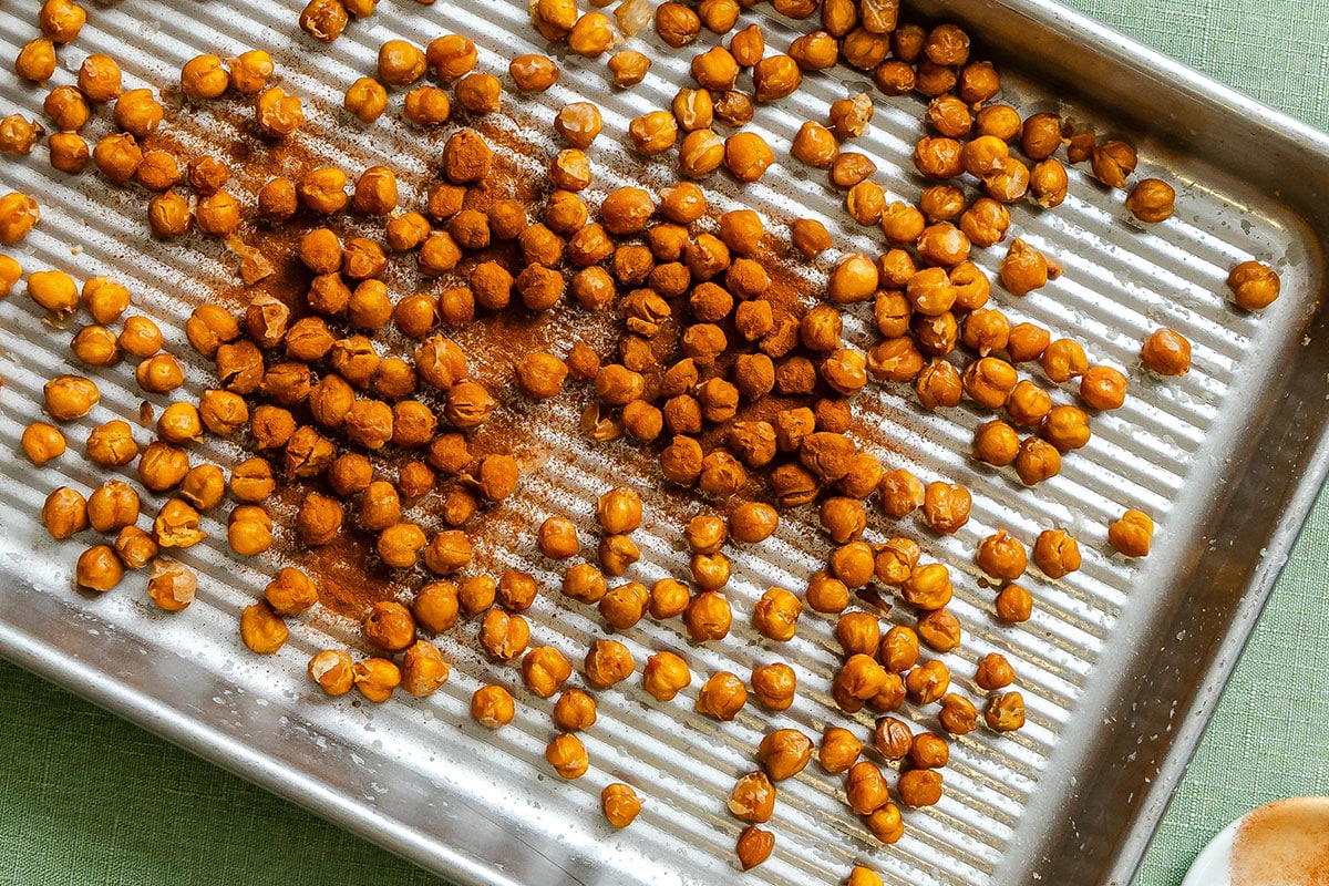 Roasted chickpeas, seasoned with spices, are spread on a ridged metal baking tray placed on a green surface.