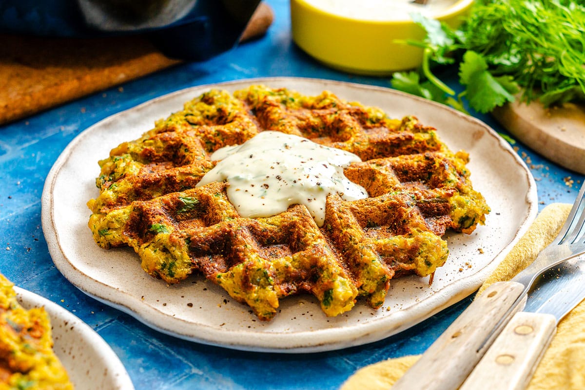 A savory waffle topped with a dollop of creamy sauce on a ceramic plate, with herbs and utensils nearby on a blue surface.