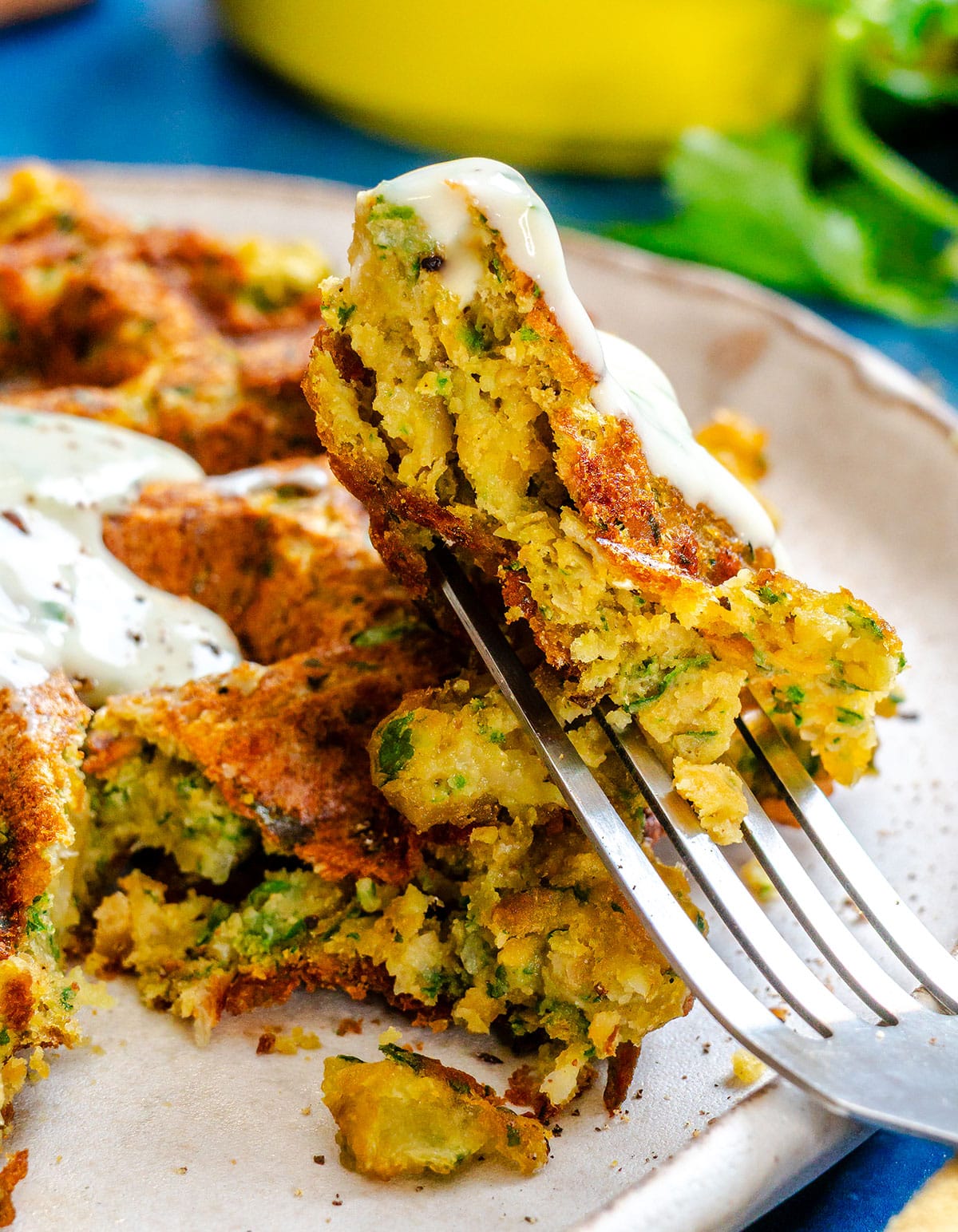 A close-up of a fork holding a piece of a golden-brown vegetable fritter topped with a drizzle of creamy white sauce on a plate.