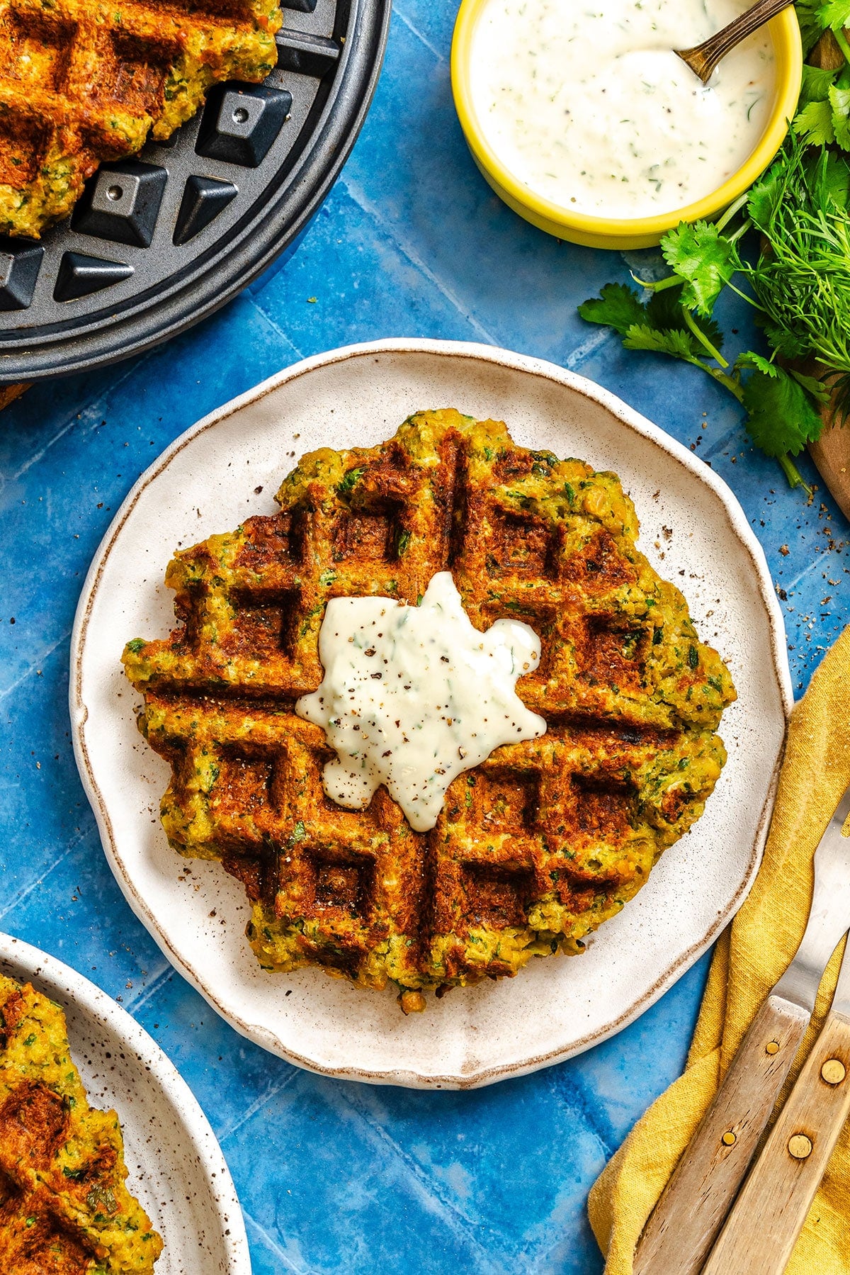 A cooked falafel waffle with green flecks sits on a plate, topped with a dollop of creamy white sauce. Nearby are a fork and knife, herbs, and a bowl of extra sauce.