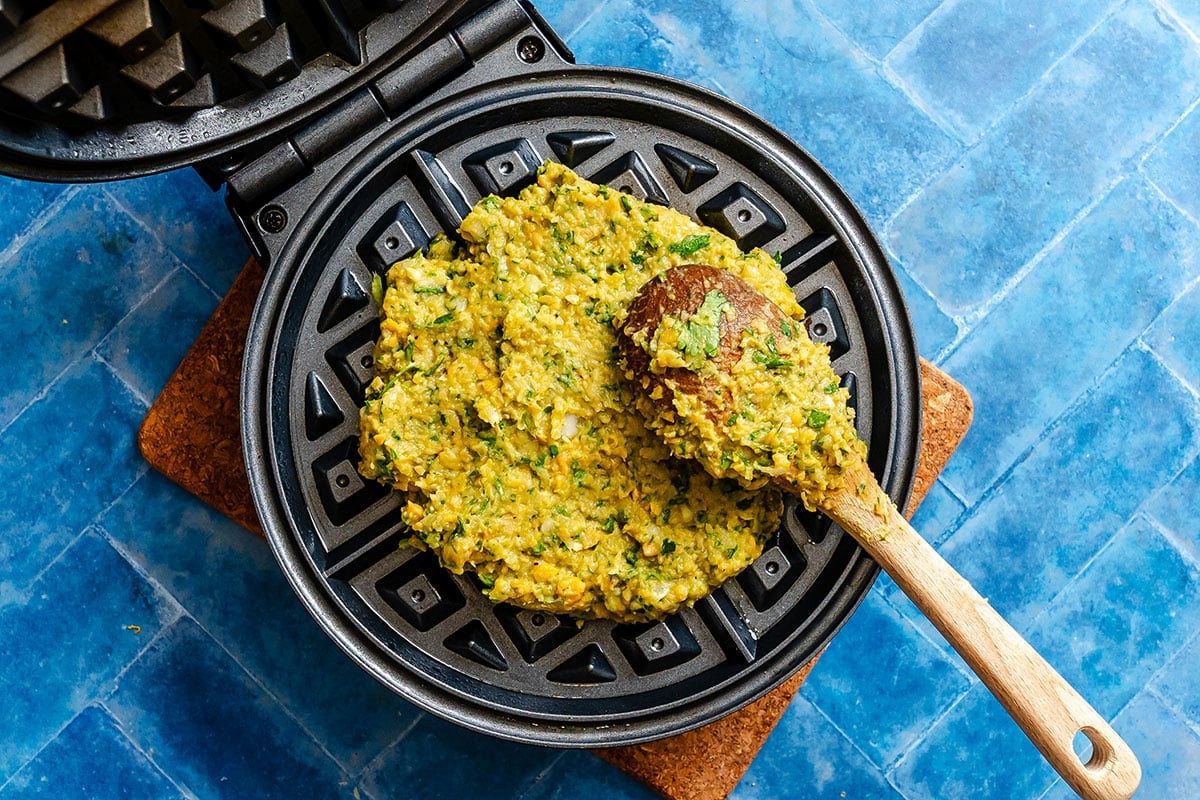 A wooden spoon spreads a thick, green and yellow batter onto a waffle maker with a blue tile surface underneath.
