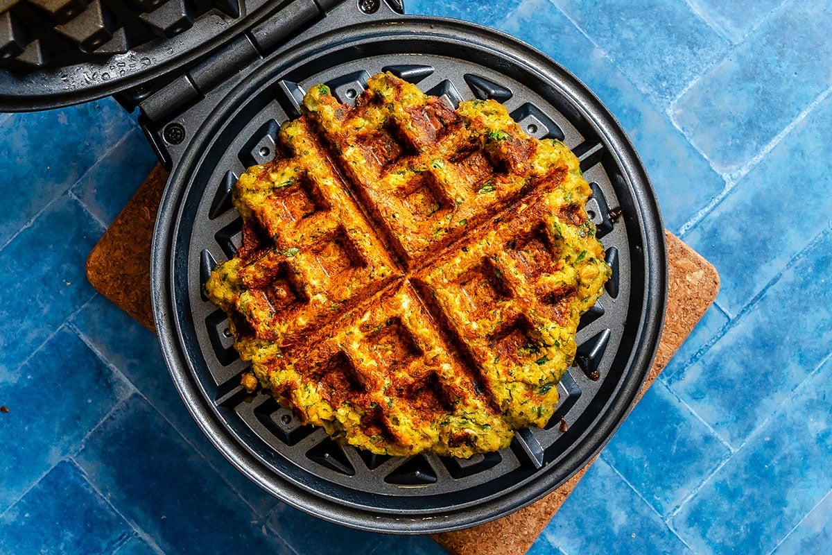 A cooked waffle with visible herbs sits inside an open waffle maker on a blue tiled surface.