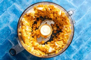 Chopped chickpeas and onions for falafel waffles in a food processor bowl, viewed from above on a blue tiled surface.