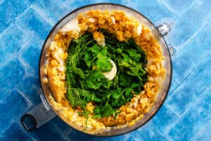 Overhead view of a food processor containing chickpeas, parsley, cilantro, and dill on a blue tiled surface.