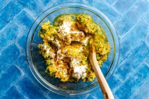 A glass bowl contains a mixture of shredded vegetables, flour, spices, and eggs, with a wooden spoon resting inside, on a blue tile surface.