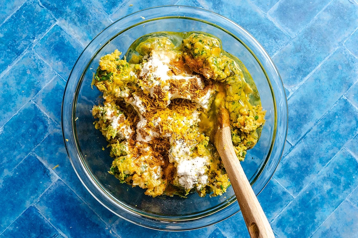 A glass bowl contains a mixture of shredded vegetables, flour, spices, and eggs, with a wooden spoon resting inside, on a blue tile surface.