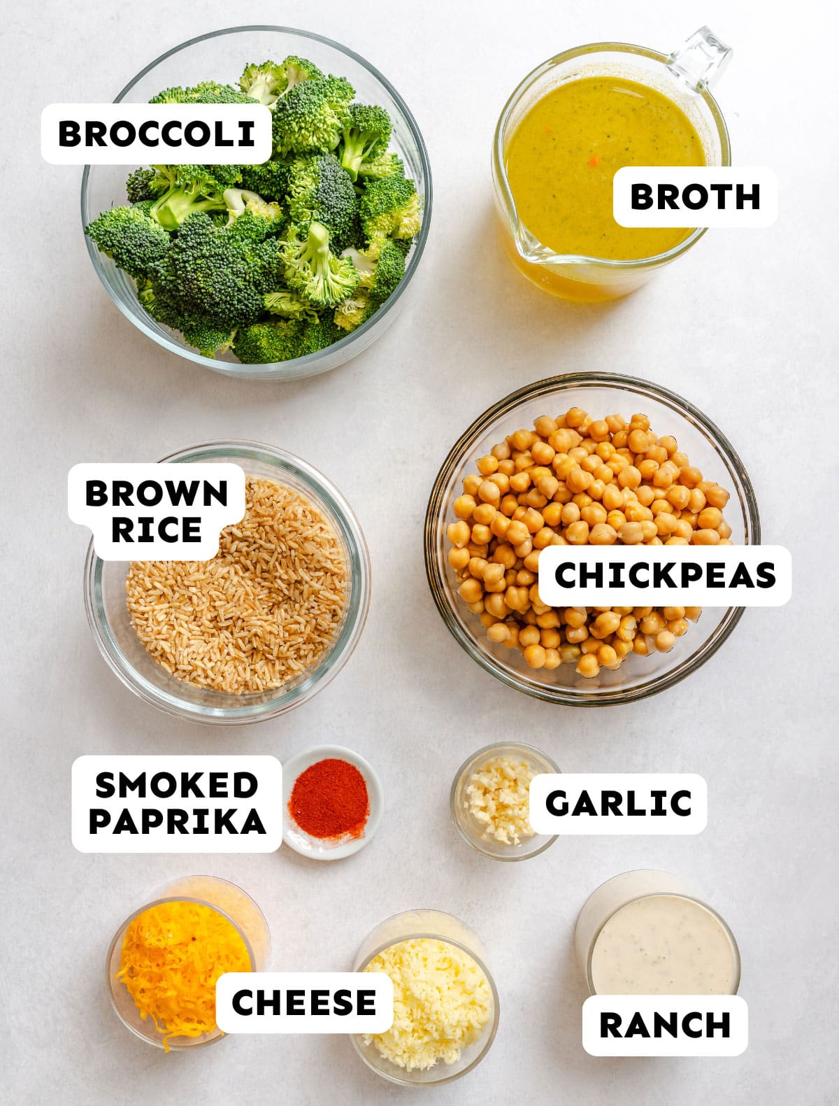 Bowls containing broccoli, broth, brown rice, chickpeas, smoked paprika, garlic, shredded cheese, and ranch dressing arranged on a white surface. Labels identify each ingredient.