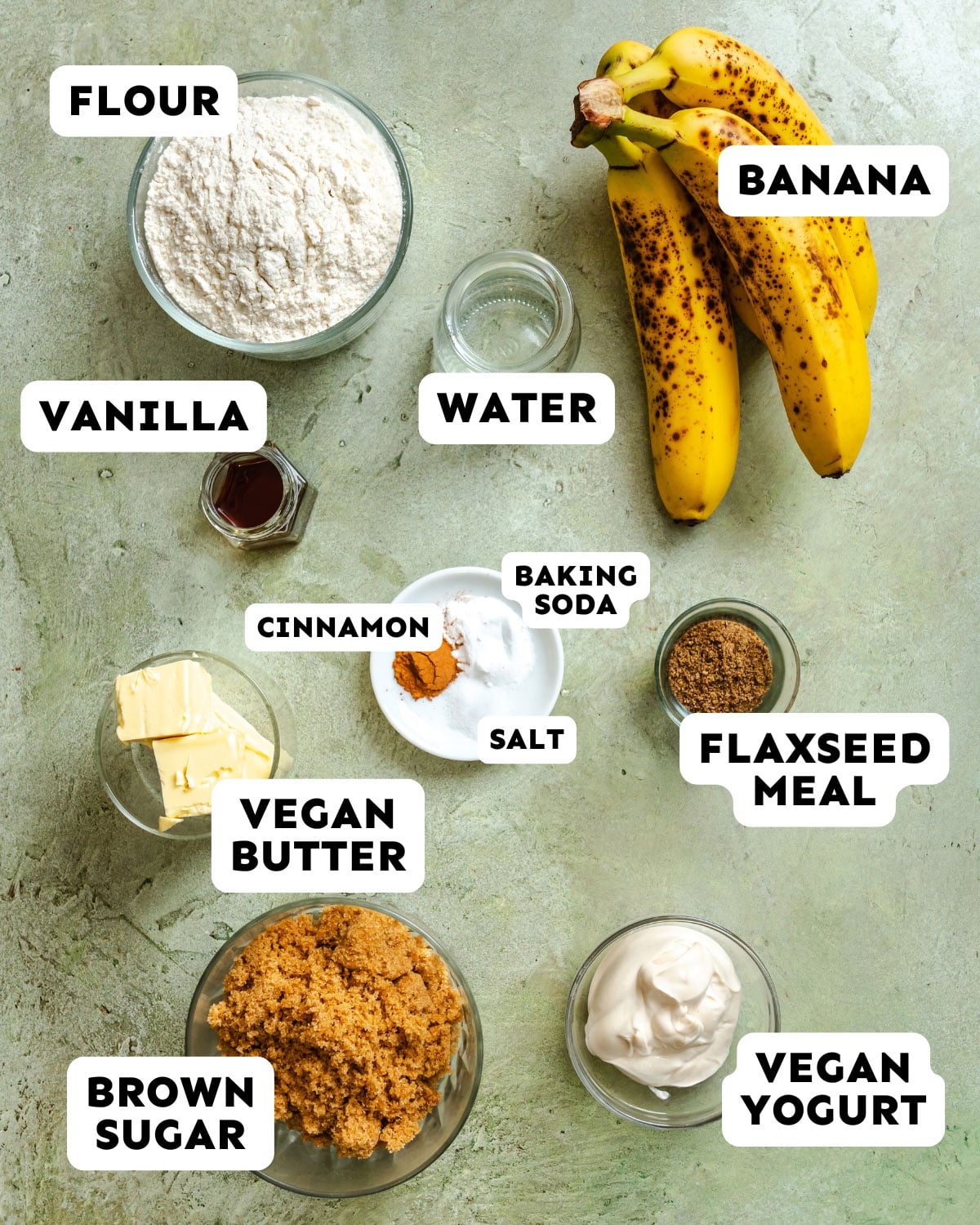 Top-down view of Vegan Banana Bread ingredients, including flour, bananas, vanilla, water, cinnamon, salt, baking soda, flaxseed meal, vegan butter, brown sugar, and vegan yogurt. Perfect for making delicious Vegan Banana Bread at home.