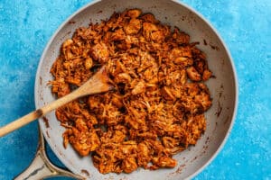 Shredded jackfruit cooked in a skillet with a wooden spoon, coated in a reddish-brown sauce on a blue background.