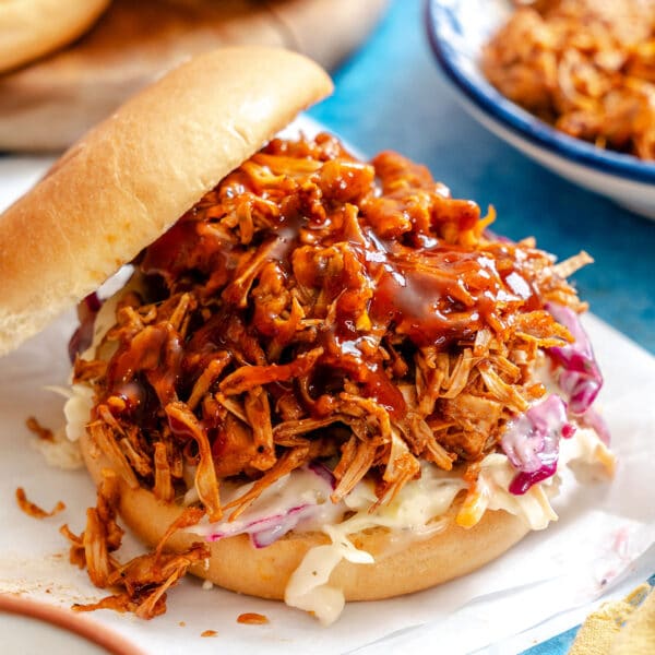 A pulled pork sandwich on a bun with barbecue sauce and coleslaw, served on a white surface with extra pulled pork and sauce in the background.