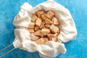 Chunks of jackfruit placed on a white cloth, with the cloth gathered inside a metal strainer, set against a blue textured background.