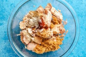 A glass bowl contains shredded jackfruit, brown sugar, and assorted spices, set on a blue surface.