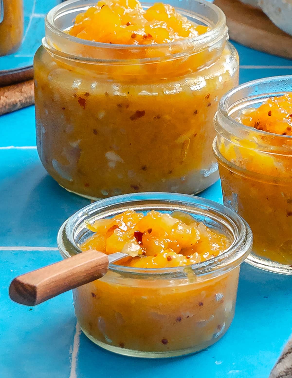 Three glass jars filled with mango chutney are placed on a blue tile surface, with a small wooden spoon resting in the front jar.