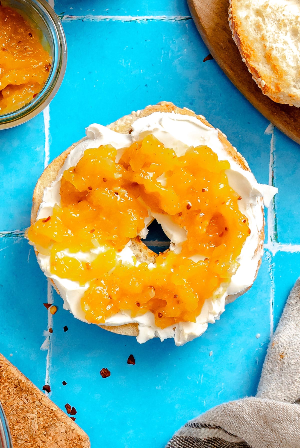 A sliced bagel topped with cream cheese and mango chutney sits on a bright blue tiled surface, surrounded by a jar of marmalade and other food items.