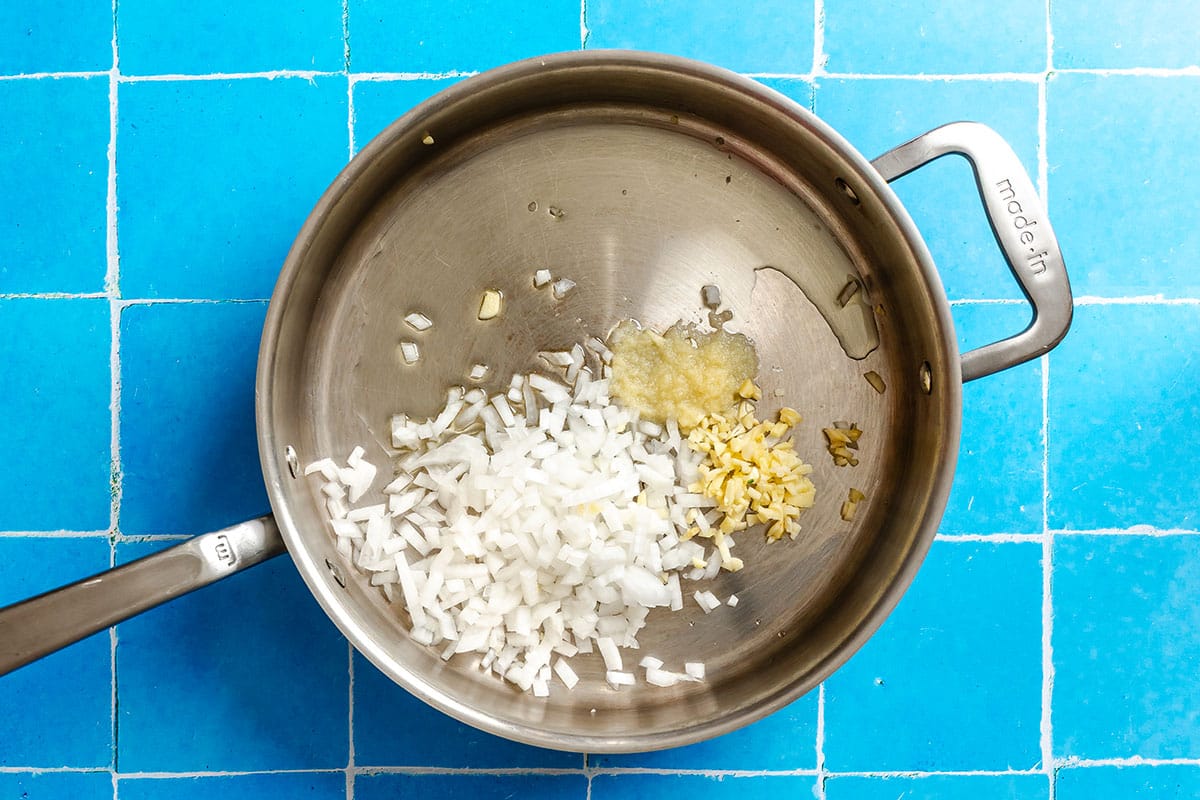 Chopped onions, minced garlic, and minced ginger in a stainless steel pan with oil, on a blue tiled surface.