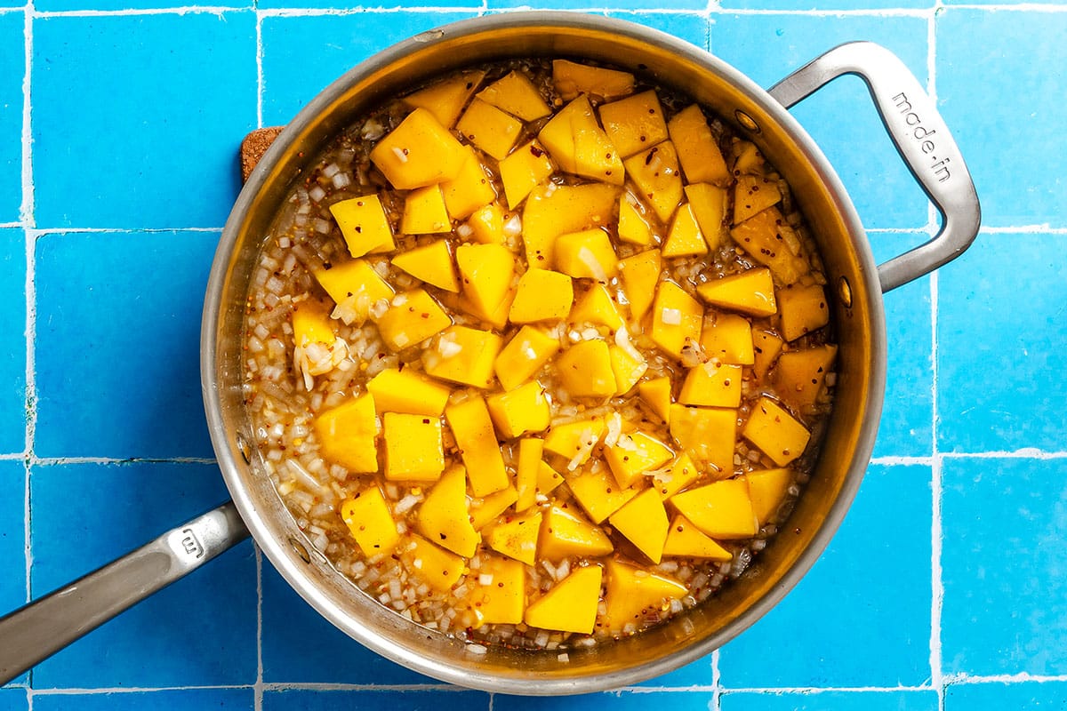 A stainless steel pot containing diced yellow mango pieces in a mixture of onions and liquid, placed on a bright blue tiled surface.