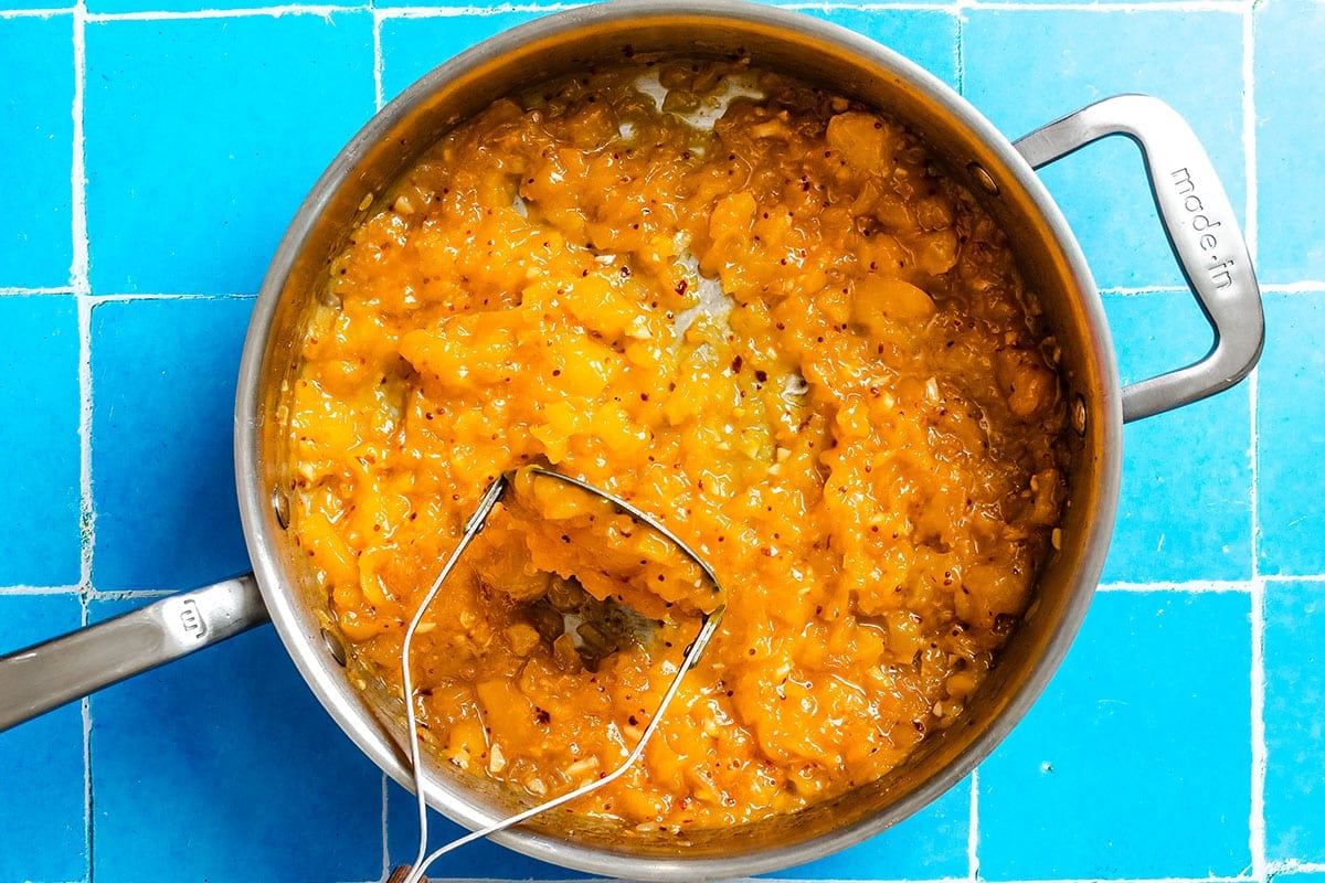 A saucepan filled with mashed mango chutney, sits on a blue tiled surface. A metal masher is partially submerged in the mixture.