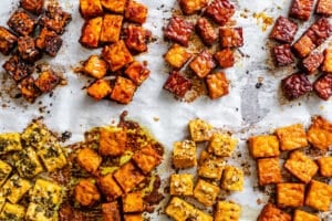 Assorted cubes of marinated and baked tofu arranged in groups on parchment paper, each group featuring a different seasoning or sauce.