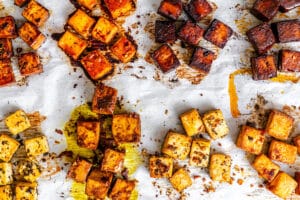 Cubed tofu pieces in various seasonings are arranged on a parchment-lined baking sheet, some with a golden, crispy texture and others darker from caramelized marinades.