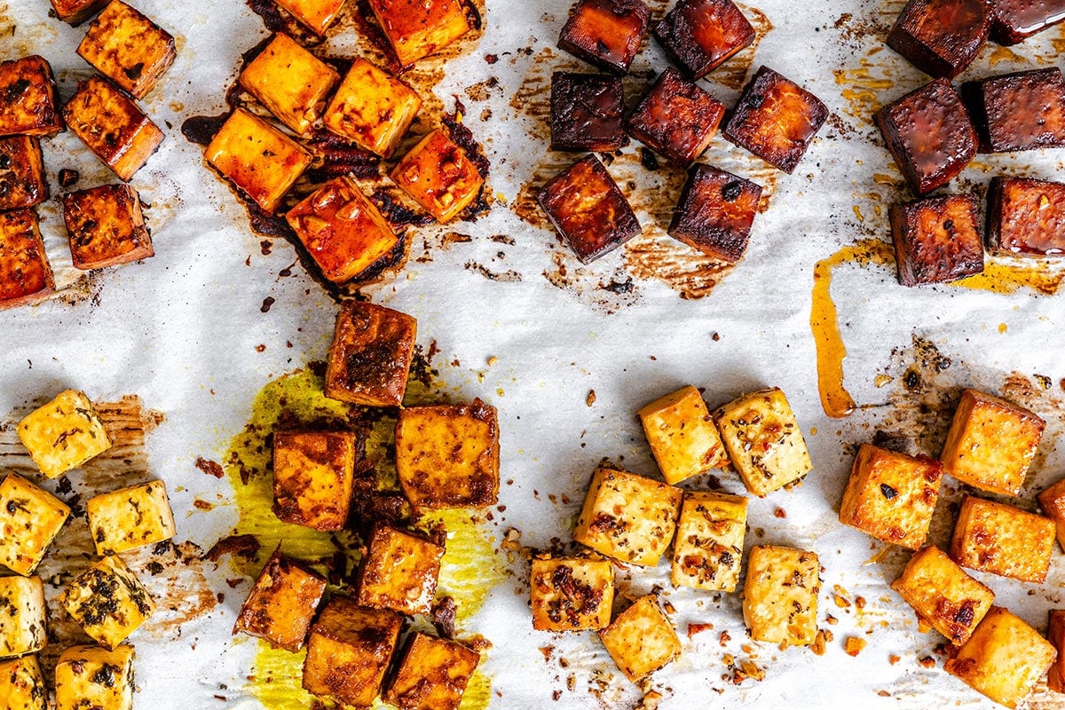 Cubed tofu pieces in various seasonings are arranged on a parchment-lined baking sheet, some with a golden, crispy texture and others darker from caramelized marinades.