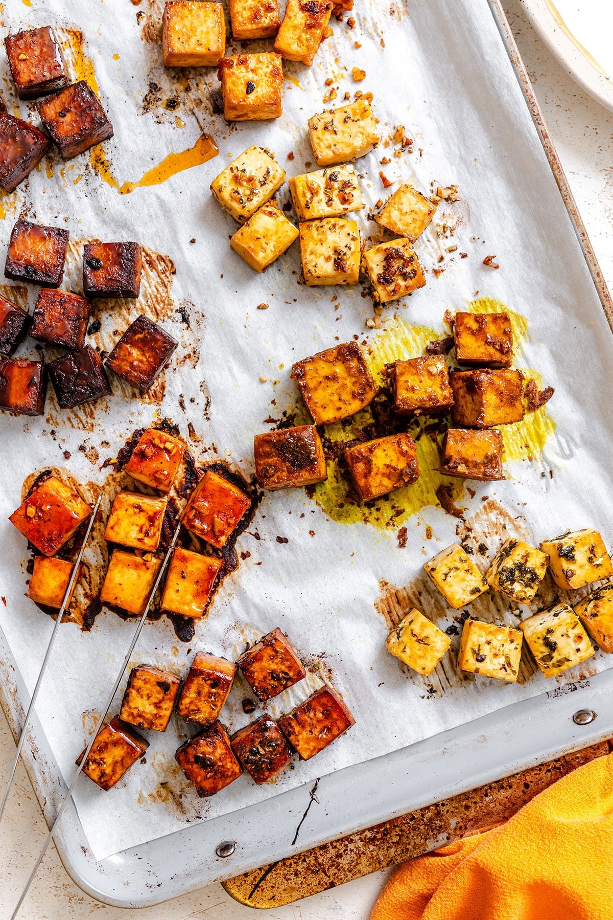 Cubes of baked tofu in various spices and marinades are arranged on a parchment-lined baking sheet.