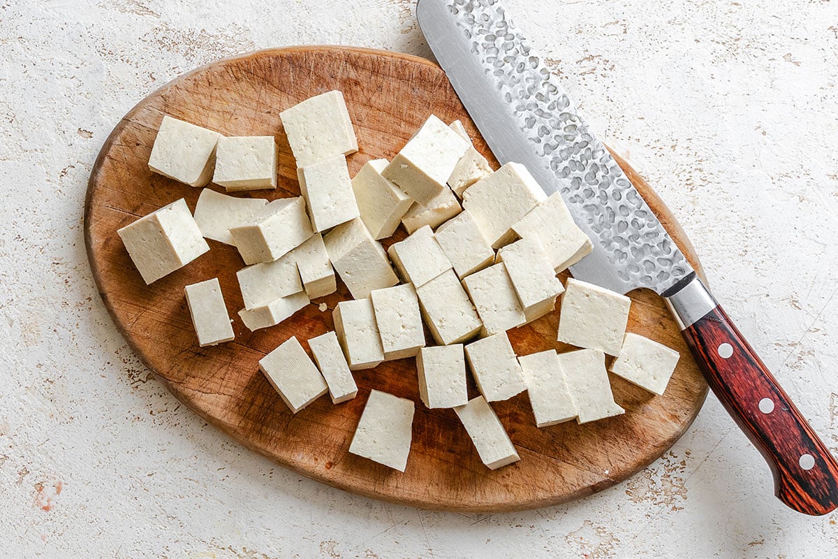 Cubed tofu on a wooden cutting board next to a large kitchen knife with a textured blade and a wooden handle.