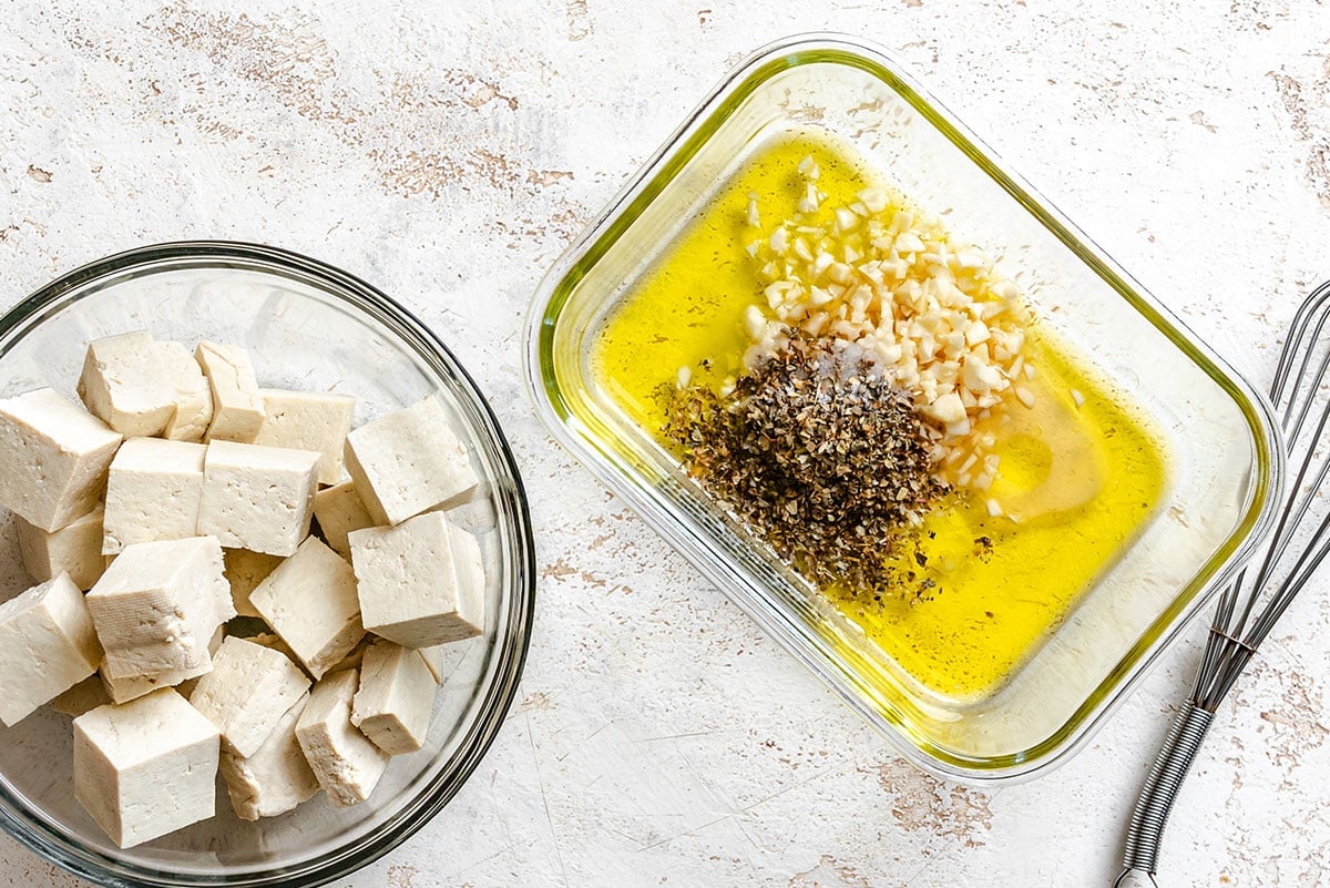 A glass bowl of cubed tofu beside a dish containing olive oil, minced garlic, and herbs on a light textured surface.