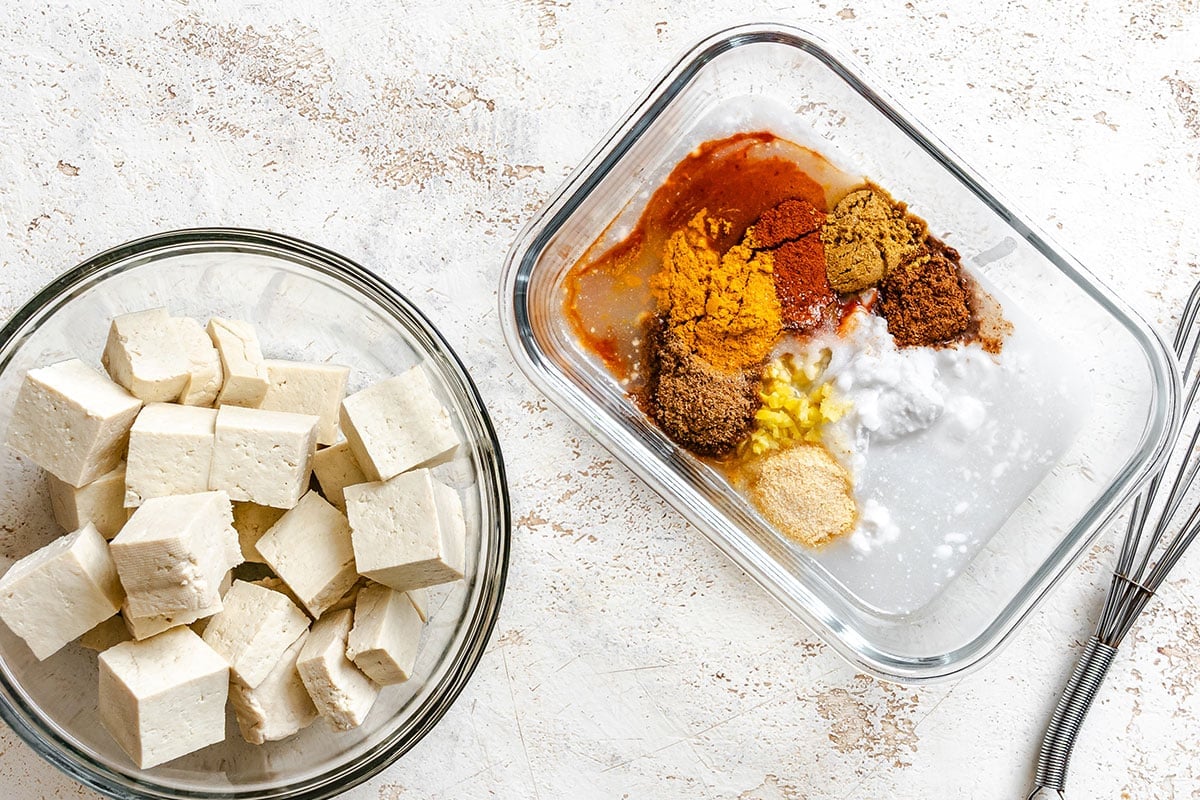 Glass bowl with cubed tofu next to a dish containing various spices and seasonings on a light textured surface.