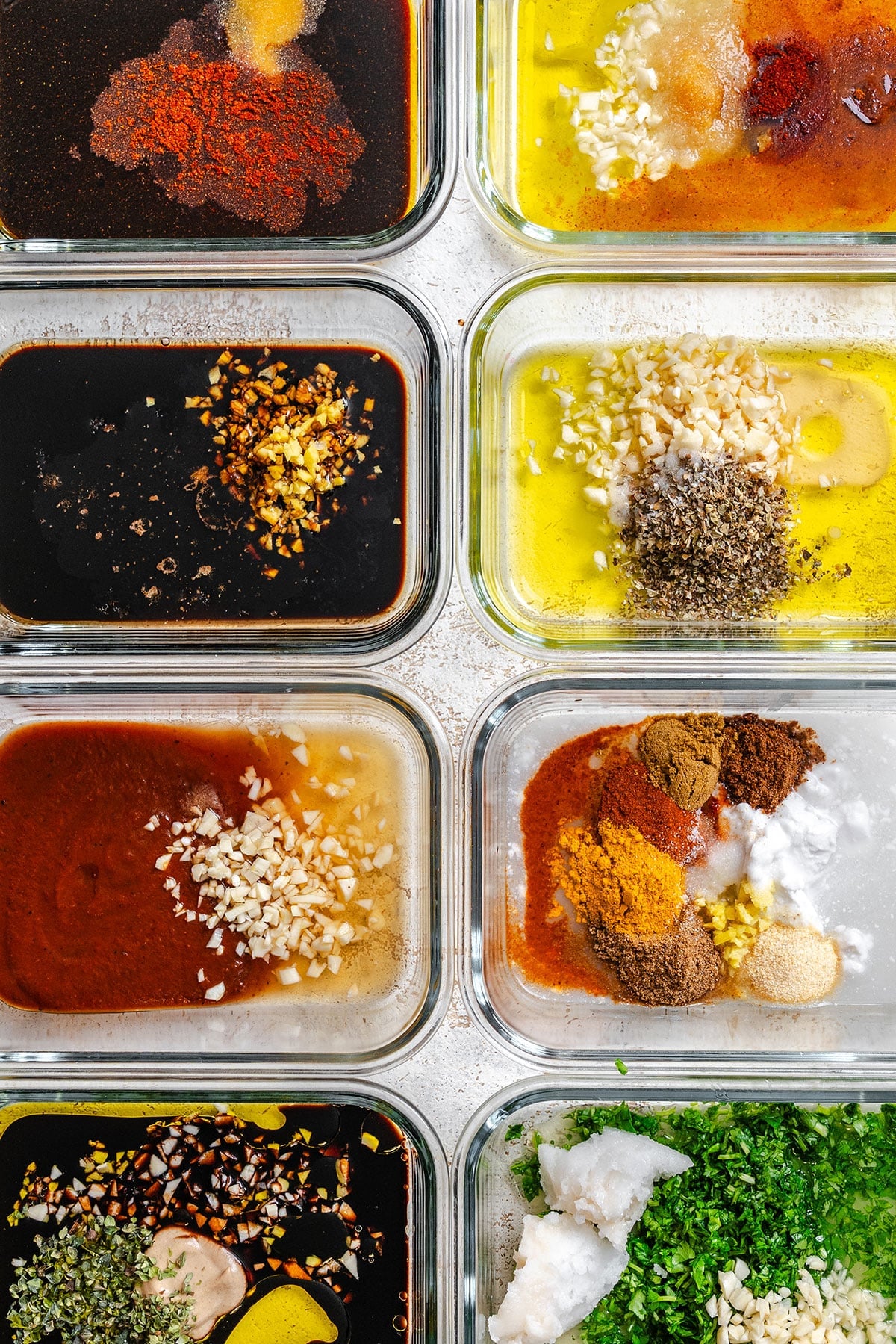 Nine glass containers filled with various marinades, spices, oils, herbs, and chopped garlic, arranged in a neat grid on a light surface.