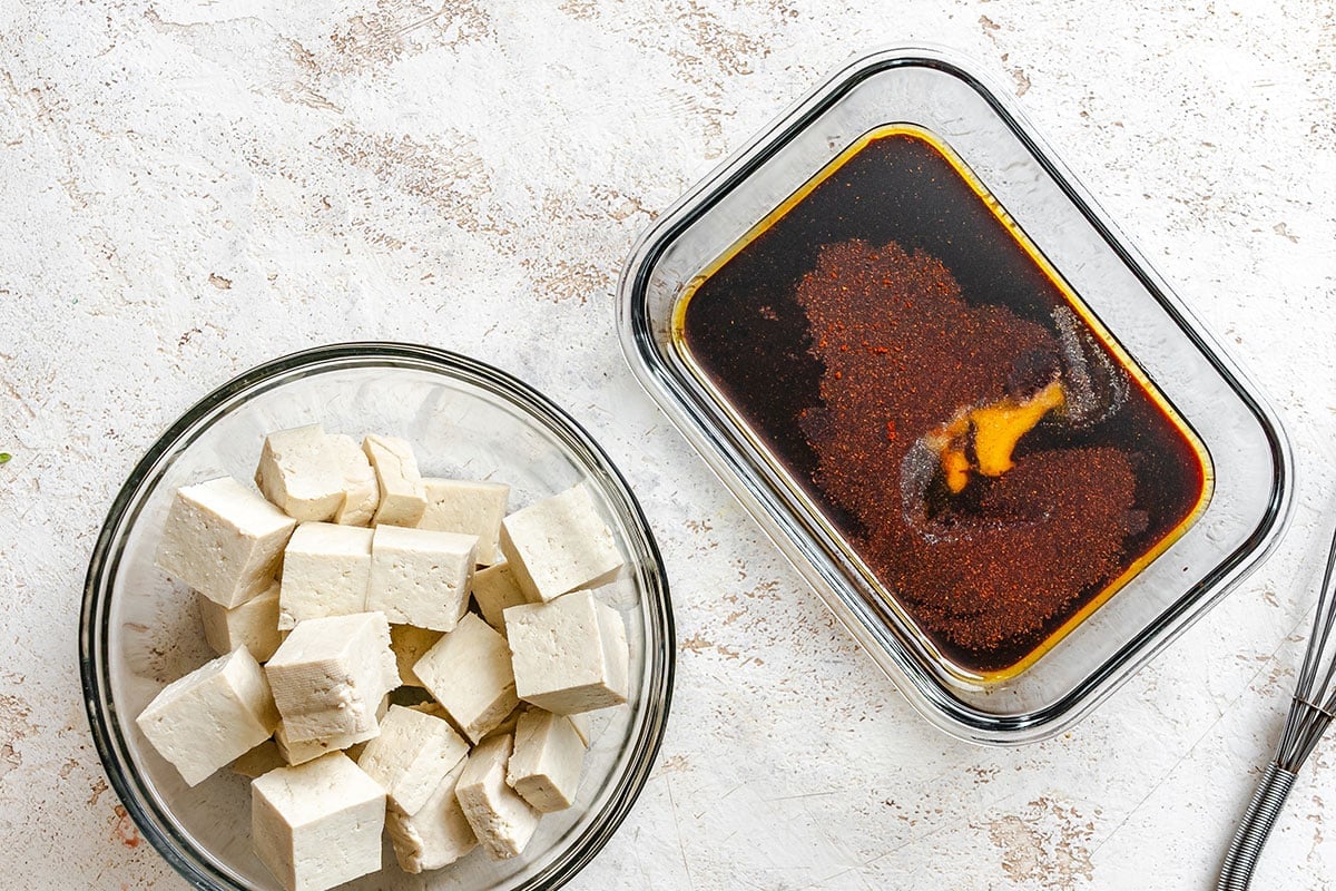 A glass bowl of cubed tofu beside a glass dish containing a dark liquid marinade on a light textured surface.