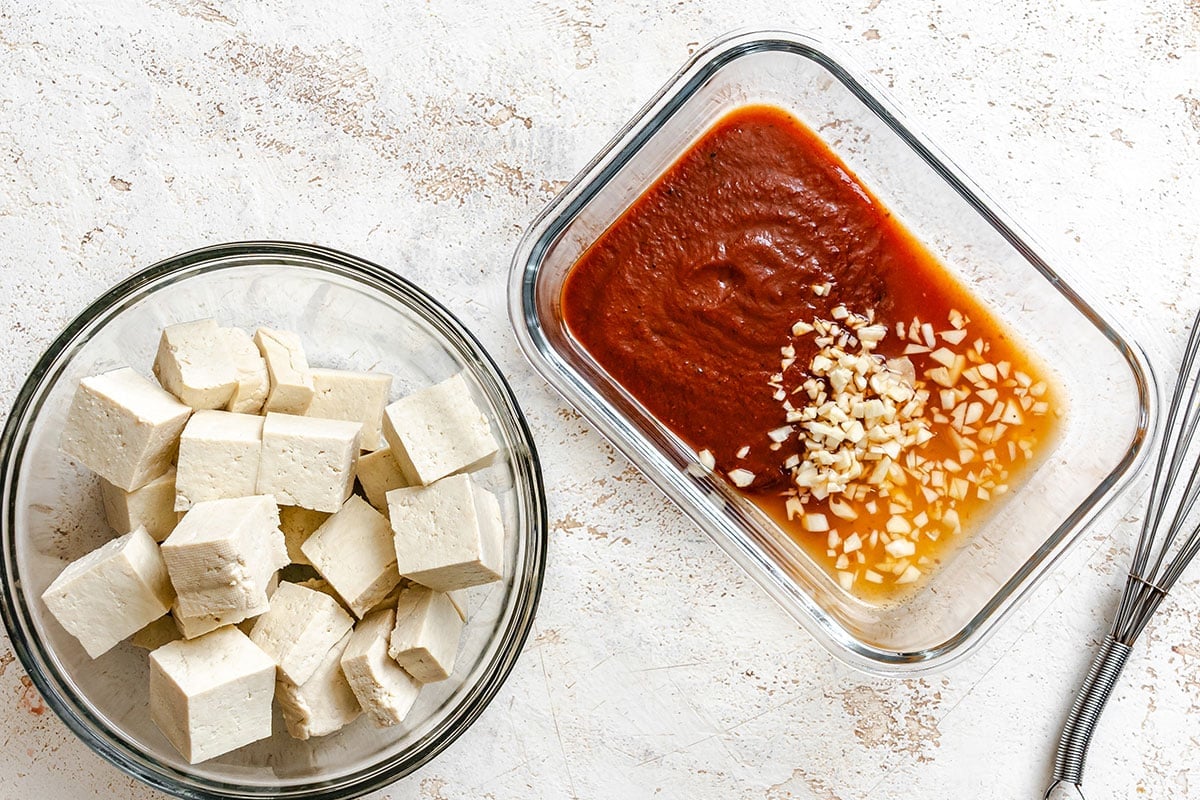 A glass bowl of cubed tofu sits next to a rectangular dish containing a red sauce topped with minced garlic and ginger.