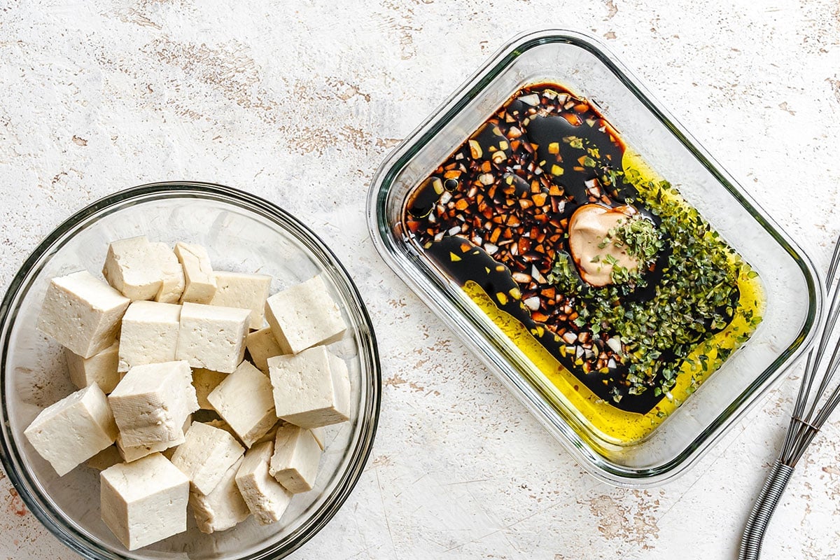 Two glass containers: one with cubed tofu, the other with a marinade of soy sauce, chopped herbs, spices, and a dollop of mustard on a light textured surface.