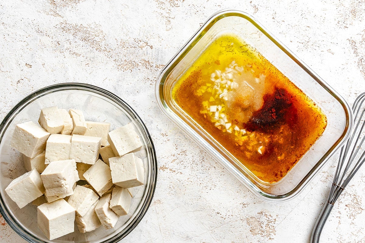 Cubed tofu in a glass bowl next to a dish containing marinade with oil, chopped garlic, and spices on a light textured surface.