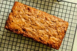 A loaf of banana bread rests on a black wire cooling rack on a light-colored surface.