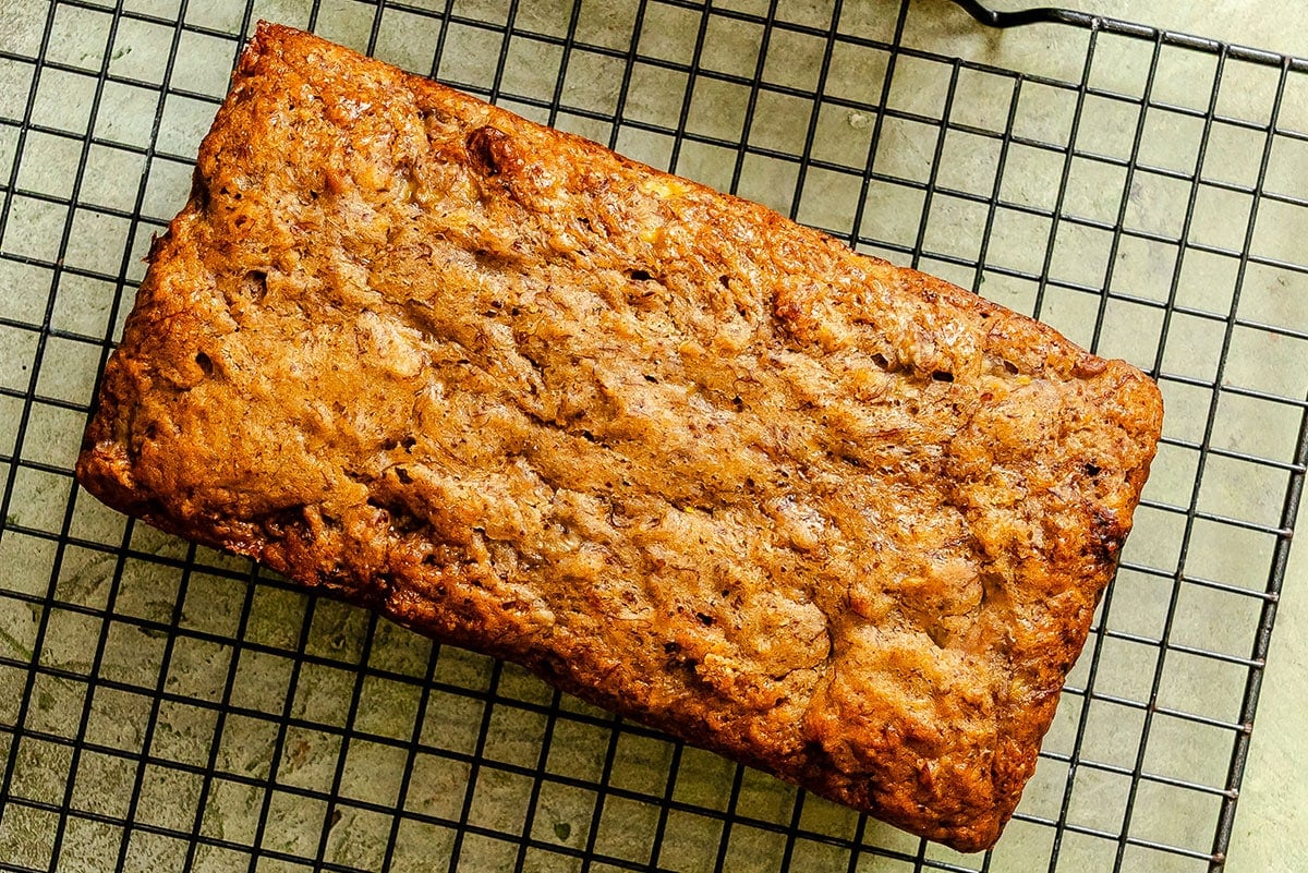 A loaf of banana bread rests on a black wire cooling rack on a light-colored surface.