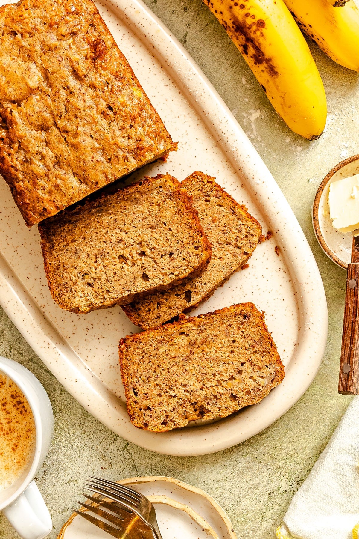 A loaf of vegan banana bread partially sliced on a plate, with two bananas, a pat of butter, a cup of coffee, and a fork nearby.