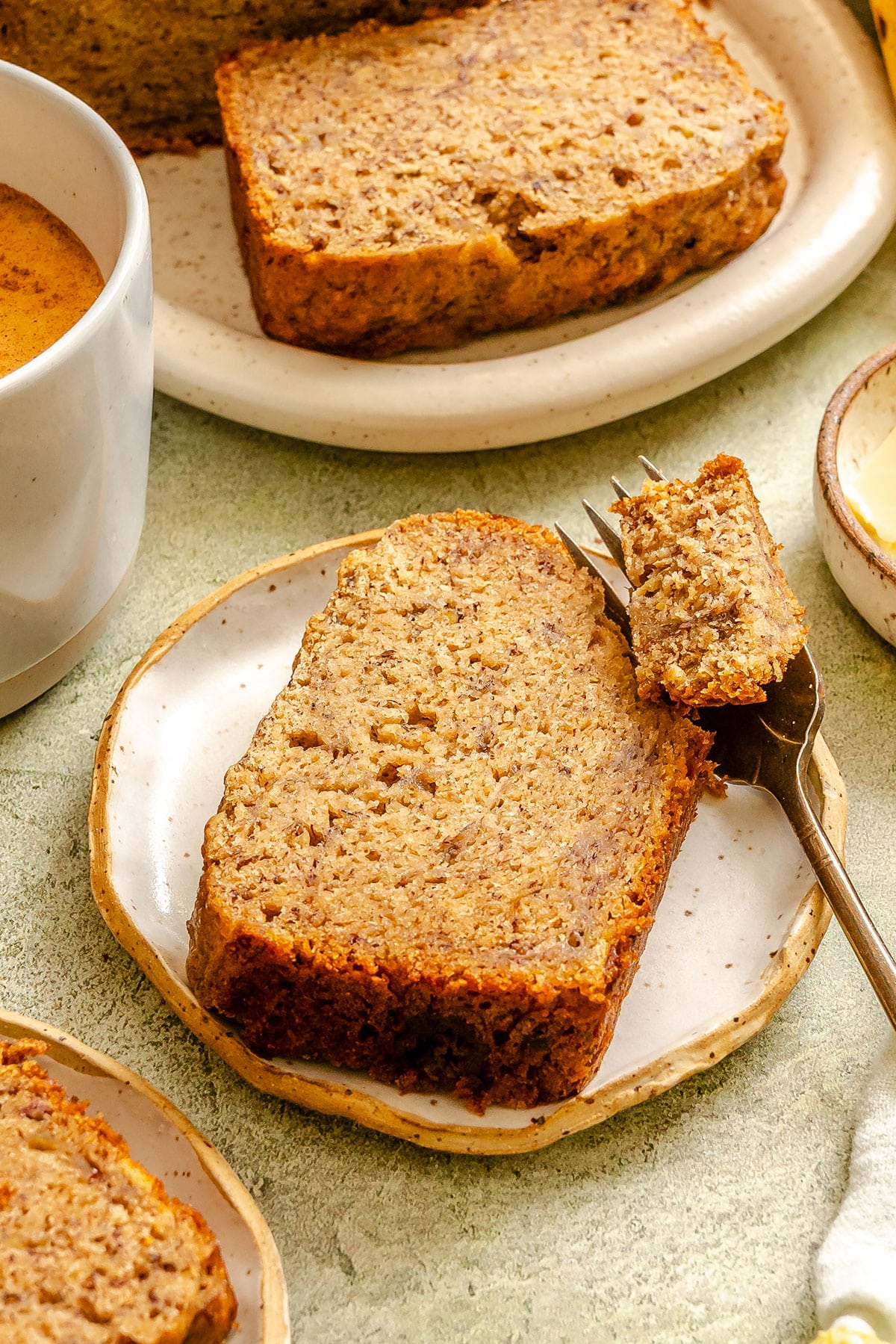 A slice of vegan banana bread on a small plate with a forkful cut out, next to a cup of coffee and another plate with more banana bread.
