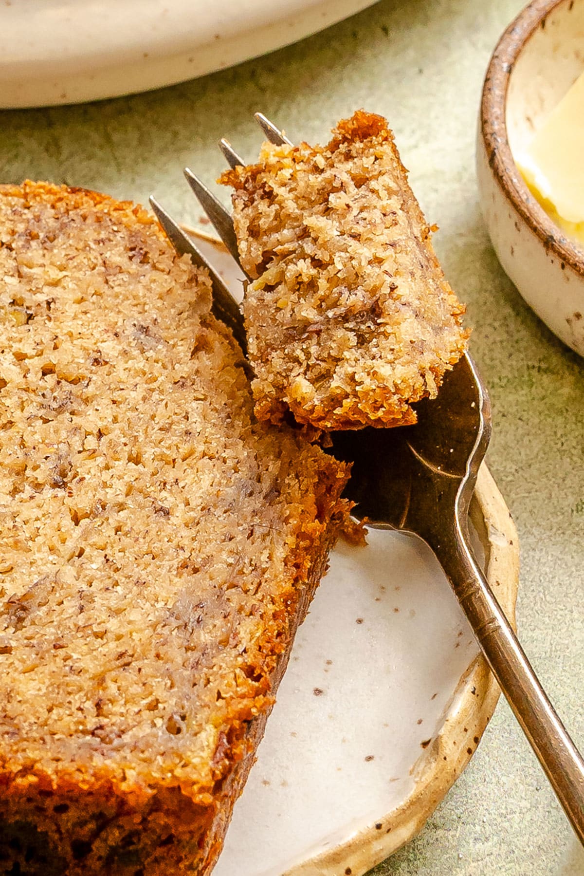 A slice of vegan banana bread on a plate with a fork holding a small bite, next to a bowl of butter.