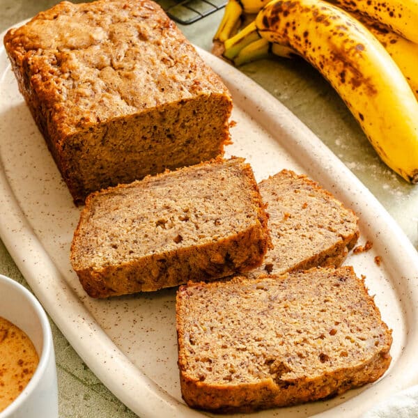 A loaf of vegan banana bread with three slices cut, served on a white platter next to a bunch of ripe bananas.