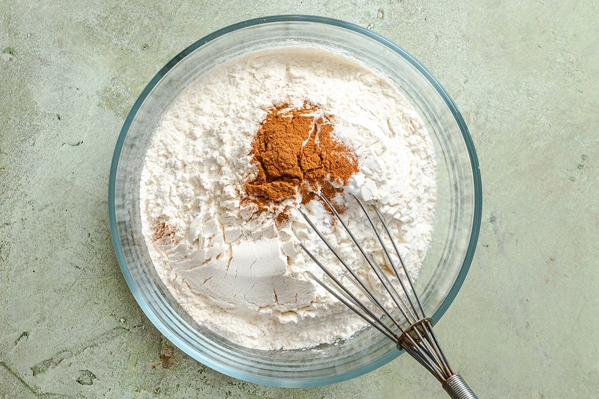 A glass bowl containing flour and ground cinnamon with a metal whisk on a light green surface.