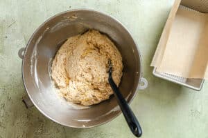 A metal mixing bowl with bread dough and a black spatula next to a bread pan lined with parchment paper on a light-colored surface.