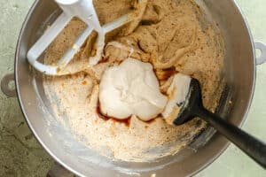 A metal mixing bowl contains a creamy brown batter, a dollop of white cream, and some vanilla extract, with a spatula and a stand mixer paddle attachment inside.