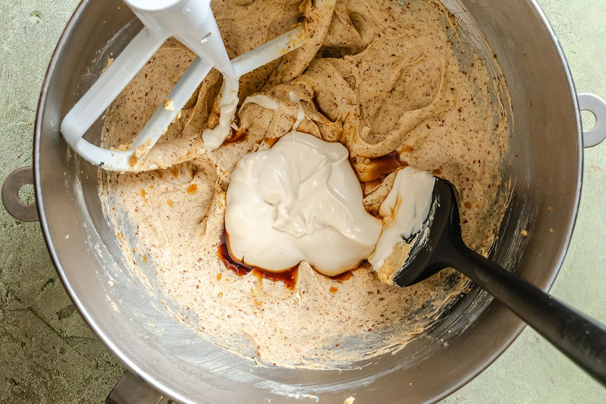 A metal mixing bowl contains a creamy brown batter, a dollop of white cream, and some vanilla extract, with a spatula and a stand mixer paddle attachment inside.