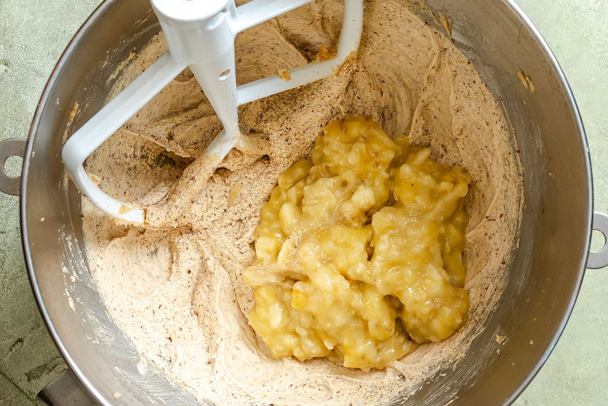Mashed bananas sit on top of partially mixed batter in a metal mixing bowl with a paddle attachment.