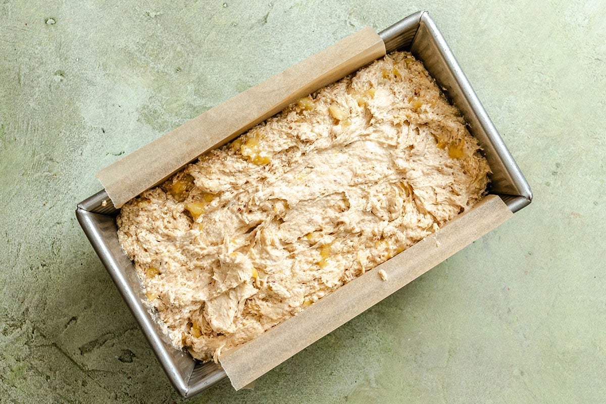 A metal loaf pan lined with parchment paper is filled with unbaked bread or cake batter, resting on a light green textured surface.